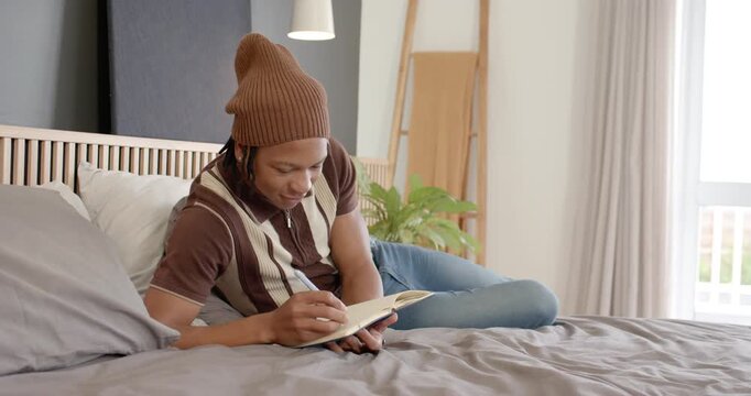 African man wearing brown beanie on bed, writing in notebook, pausing to think, gazing at window