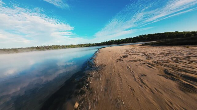 Aerial view of misty lake at dawn. Water reflects blue sky and clouds. Sandy shore meets calm water. Forest borders lake's edge. Mist rises gently over surface. Serene, peaceful natural scenery