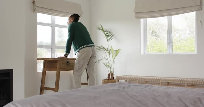 Adult African man leaning at wooden desk in bedroom, noticing view outside and adjusting blind
