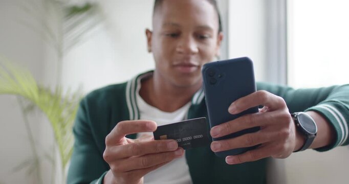 African American man in cardigan on sofa aligning card with phone scanning chip confirming payment