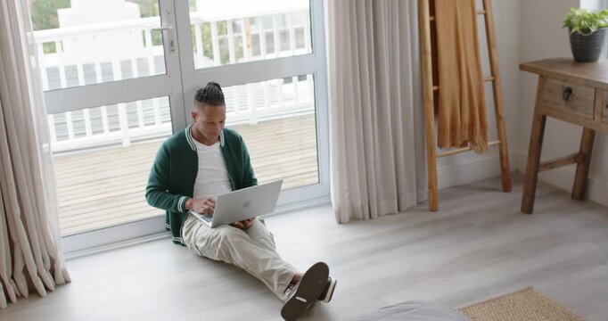 African American man opening laptop and typing on lap by sliding glass door, working remotely