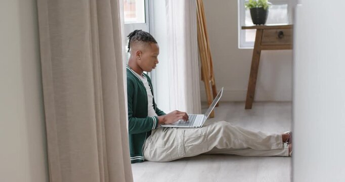 African American man placing laptop on lap, typing, working, glancing at window, in green cardigan