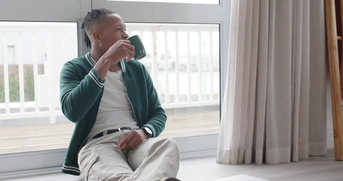 African American man sitting by sliding glass door holding green mug, sipping while gazing outside