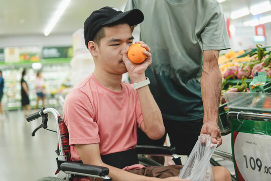 Young Asian man in a wheelchair smelling a fresh orange to check freshness while grocery shopping with an assistant at the supermarket, concept of inclusive lifestyle and accessibility.