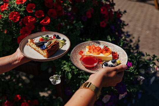 Hands hold two plated cake desserts with berries, cream, fruit, edible flowers and red sauce beside vibrant blooming flowers outdoors