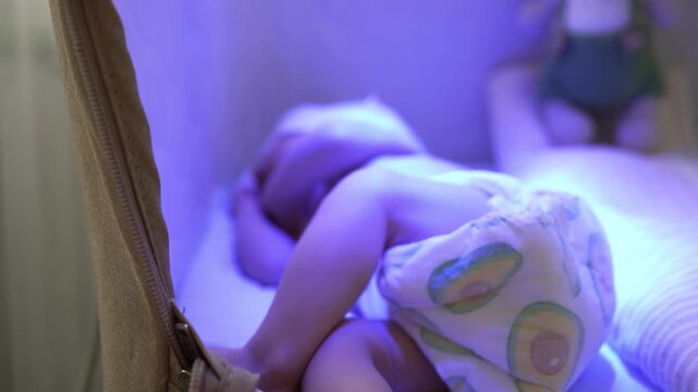 Tiny newborn baby with jaundice sleeping in a bassinet under a special blue ultraviolet light for phototherapy, a common home treatment to reduce high bilirubin levels