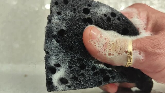 A woman's hand squeezes and soaps a black dishwashing sponge, creating foam while preparing to wash dishes, household cleaning and kitchen routine.