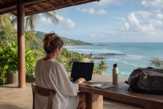 Female Digital Nomad Working on a Laptop from a Shaded Tropical Terrace overlooking a Peaceful Ocean Coastline during a Flexible Workday