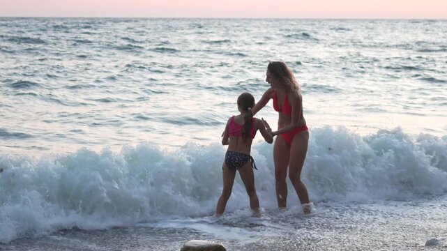 Playing mother and daughter hold hands and splash in coastal sea surf at sunset. Medium tracking shot.