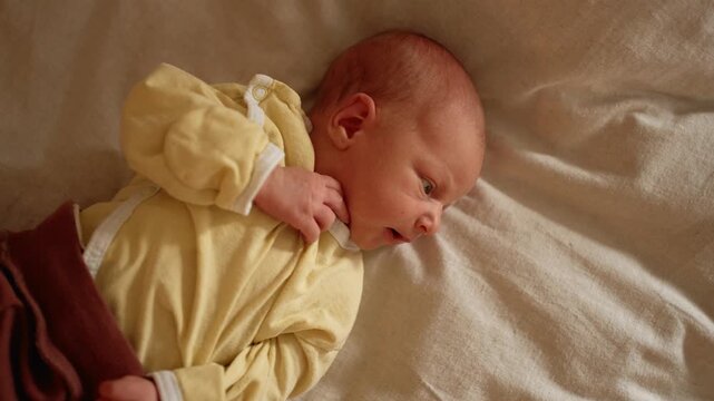 Newborn baby boy with big curious eyes making funny faces and grimaces while lying on a bed, expressing different emotions as the camera gets closer for an intimate portrait