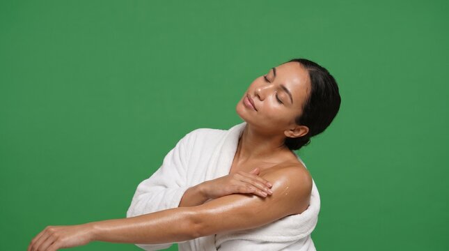 Hispanic young adult woman in Generation Z wearing a white plush bathrobe, resting her hand on her bare shoulder with head tilted back against a solid green screen background.