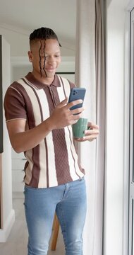 Vertical video: Man standing near window checking smartphone messages while sipping from green mug