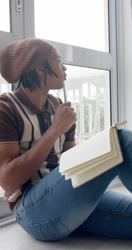Vertical video: African American man sitting at window gazing, writing notes in notebook using pen