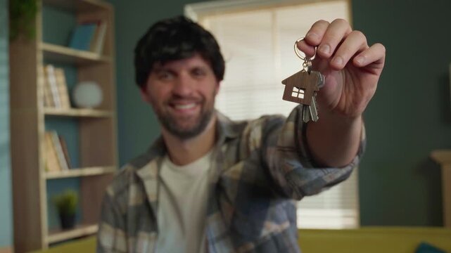 Happy bearded man holding up a keychain with house shaped trinket and keys, celebrating buying a new apartment and showing his success and happiness after signing the real estate contract