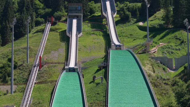 Aerial view of the ski jumping ramps at predazzo ski jumping stadium in the dolomites, showing the large and normal hills surrounded by lush alpine forest in summer.