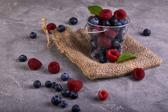 Fresh blueberries and raspberries in a transparent bucket on a gray concrete background