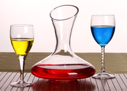 Composition of 3 items, a decanter and glasses with multi-colored liquids. Red, blue and yellow on a white background.