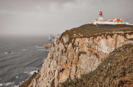 Lighthouse at Cape Roca (Cabo da Roca), most western point of Europe at coast of Atlantic Ocean in Portugal. Rocks at coastline and blue sky with clouds over skyline. Picturesque summer landscape.
