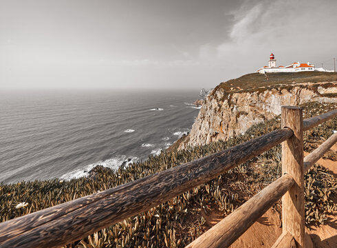 Lighthouse at Cape Roca (Cabo da Roca), most western point of Europe at coast of Atlantic Ocean in Portugal. Wooden fence, rocks at coastline and blue sky with clouds over skyline. Summer landscape.
