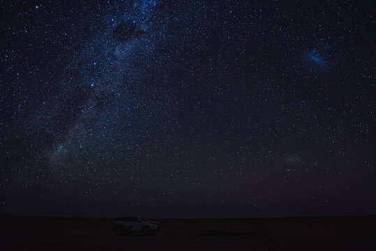 The Milky Way galaxy, Large Magellanic Cloud, Small Magellanic Cloud and constellations shine over a safari vehicle parked on the dark horizon in the desert landscape of Namibia.