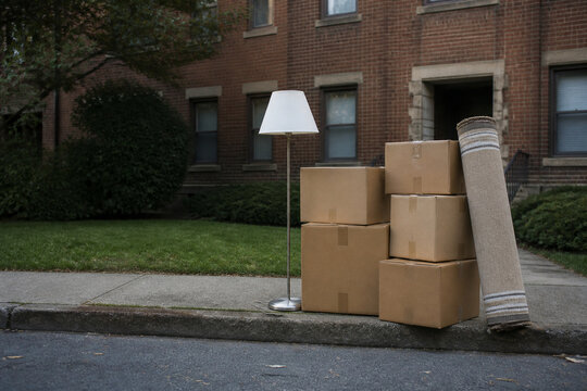 Cardboard moving boxes, rolled rug and standing lamp on curb outside brick apartment house, eviction and moving day concept