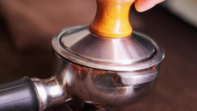 Close-up of a barista's hands pressing ground coffee with a wooden-handle tamper into a stainless steel portafilter