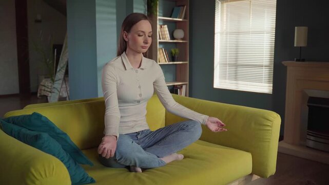 Young spiritual woman sitting in a lotus position on a sofa, peacefully meditating with closed eyes before starting to levitate and float in the air in her cozy living room at home