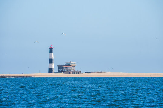 Pelican Point lighthouse and lodge stand on the sandy peninsula near Walvis Bay across the ocean water under a clear daylight sky in Namibia.