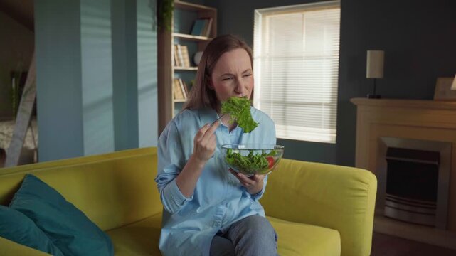 Young woman on a diet, forcing herself to eat a tasteless vegetable salad with a look of disgust while sitting on a yellow sofa at home, feeling unhappy with her healthy lifestyle choice