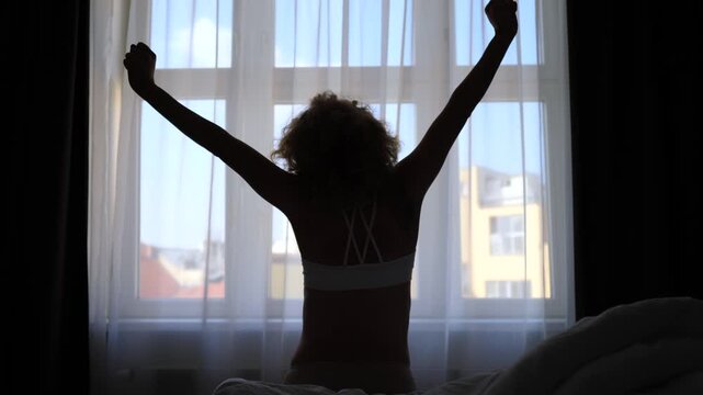 Lovely woman with curly hair stretching her arms after waking up, sitting on a bed in a dark room. Silhouette of her body against a bright window starting a beautiful new day at home