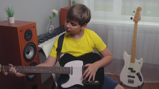 Young boy in yellow shirt playing black electric guitar during music lesson at home. Caucasian child practicing musical instrument in cozy room with speakers keyboard synth and bass guitar. 