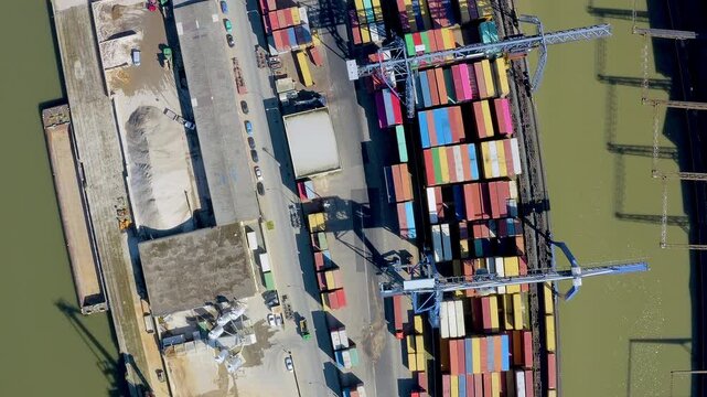 Top down aerial drone view of the Osthafen container terminal quay with a gantry crane, stacked shipping containers, silos, sand piles and a barge, Frankfurt, Germany.