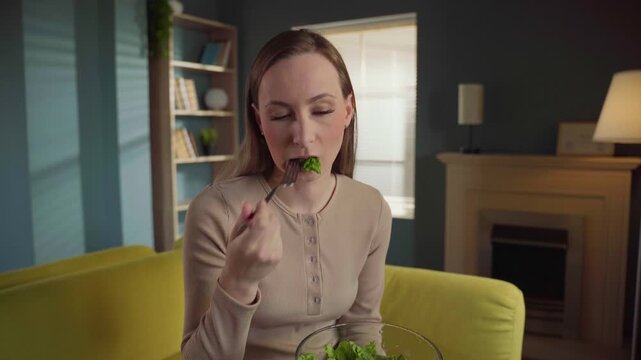 Unhappy young woman on a strict diet, feeling sick and disgusted while reluctantly eating a tasteless green salad for lunch in her living room, forcing herself to follow a healthy lifestyle