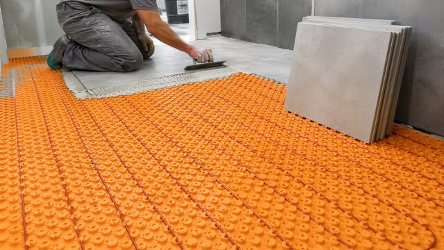 Man tiler installing ceramic tile over electric underfloor heating mat with orange uncoupling membrane and cable. Warm floor system renovation and interior construction technology concept