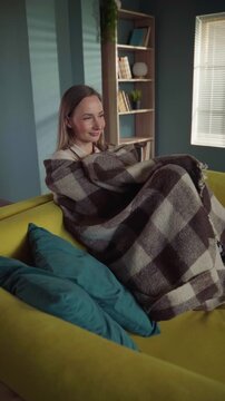 Peaceful young woman wrapped in a warm plaid blanket, resting and relaxing on a comfortable yellow sofa in her cozy living room, enjoying a quiet moment of tranquility and comfort at home