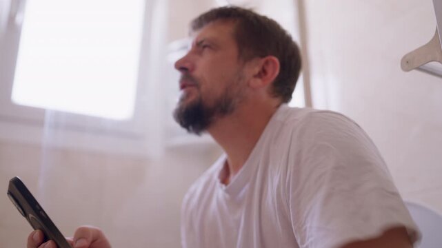 Middle-aged man absorbed in smartphone while sitting on toilet, displaying a satisfied facial expression during private leisure time, illustrating digital habits, personal space and modern lifestyle