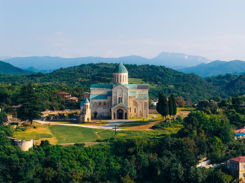 Aerial view of the historic Bagrati Cathedral with its distinctive green dome and stone architecture, surrounded by lush green hills and mountains in Kutaisi, Imereti, Georgia.