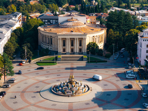 Aerial view of the Meskhishvili Theatre and the ornate Colchis Fountain in the central square, surrounded by historic buildings, lush trees, and urban streets in Kutaisi, Imereti, Georgia.