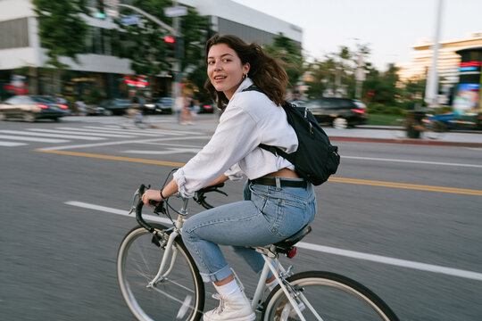 Smiling young woman with a backpack rides a bicycle on a busy urban street with motion blur, looking back at the camera, embodying an active, healthy, and eco-friendly city lifestyle during