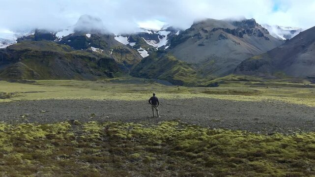 Solitary explorer standing in a vast, remote valley in Iceland, contemplating the dramatic, snow capped mountains and green landscape