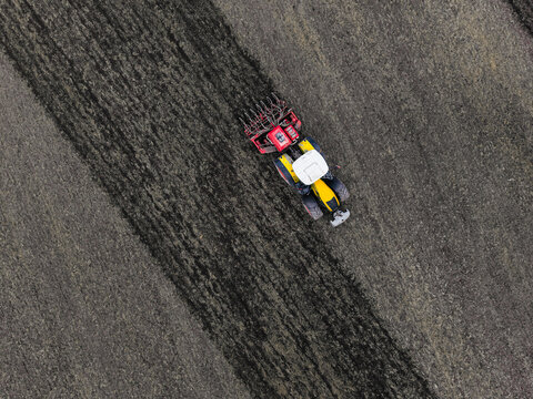 Aerial view of a yellow tractor pulling a red agricultural implement across a dark tilled field in Latvia.