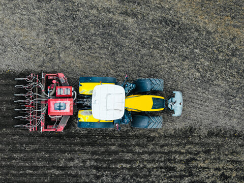 Aerial view of a yellow and black tractor pulling a seed drill across a dark, tilled agricultural field in Latvia.