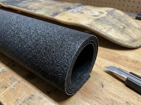 Roll of black skateboard grip tape and a utility knife on a wooden workbench for deck maintenance and gear application