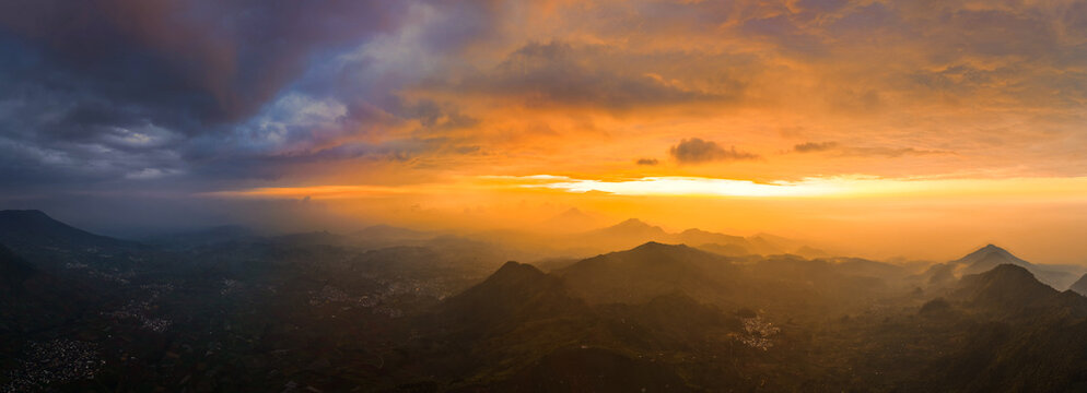 Aerial view of Telaga Dringo surrounded by dramatic mountain peaks and lush valleys under a vibrant golden sunset sky in Indonesia.