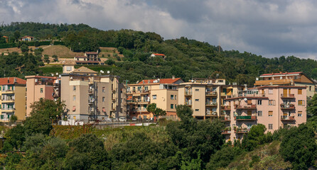 Obraz premium Vista panorámica de un pueblo mediterráneo con coloridos edificios residenciales en la ladera de una colina verde bajo un cielo nublado.