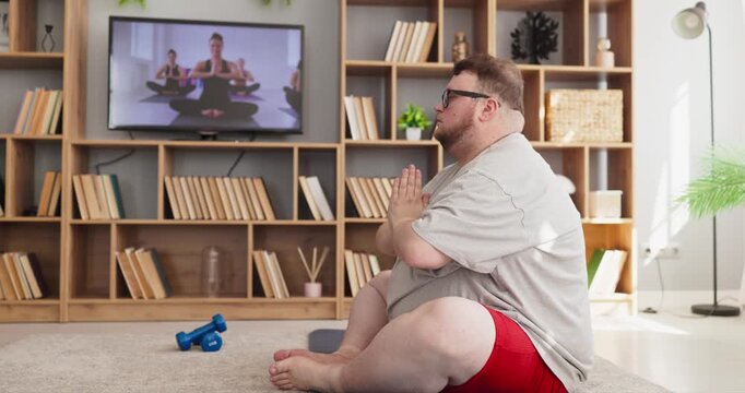 Man overweight yoga home fitness workout online training. He sits in meditation on a mat, streaming a class on TV and doing breathing for balance and calm. Calm home wellness concept today.