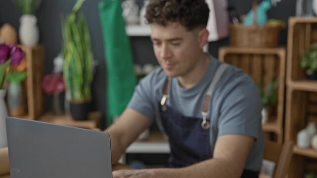 Young florist man typing on laptop with hands on keyboard among flowers and vases in studio; focused craft business entrepreneurship.