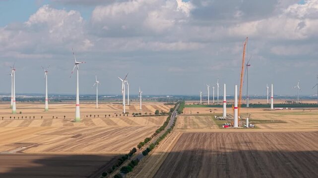 Aerial panoramic drone view wind farm expansion across harvested farmland, with crane turbine tower rising under dramatic clouds. Renewable energy development transforms rural landscape clean power