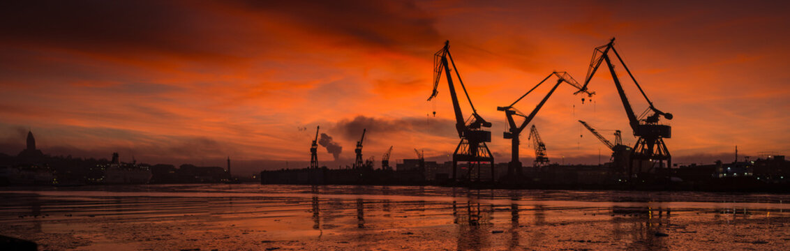 Wide panoramic view of harbor cranes at sunset with glowing sky reflection.