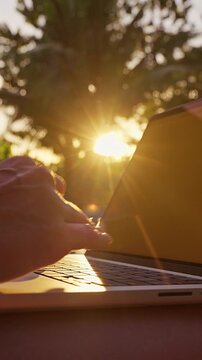 Vertical video. Close-up of hands typing on a laptop outdoors during sunset near a tropical swimming pool. Golden sunlight illuminates the keyboard, symbolizing remote work, online communication, digi